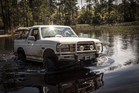 1995 Ford Bronco - A white 1995 Ford Bronco navigates through shallow water, its brush guard equipped for off-road adventure.