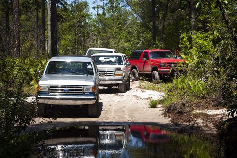 1995 Ford Bronco - Multiple Ford Broncos navigate a wooded dirt path, with reflections visible in the water ahead.