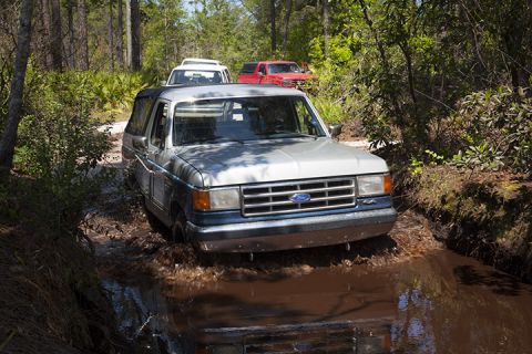 1995 Ford Bronco - A Ford Bronco navigates a muddy path in a wooded area, with other vehicles following.