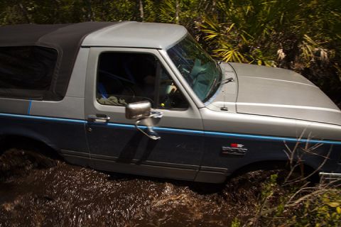 1995 Ford Bronco - A two-tone Ford Bronco navigates through a muddy off-road trail with dense foliage surrounding it.