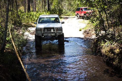 1995 Ford Bronco - Two off-road vehicles navigate a water crossing in a wooded area.