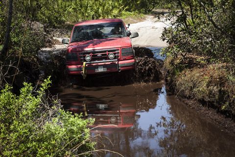 1995 Ford Bronco - 1995 Ford Bronco navigates a challenging muddy trail, splashing water and mud.