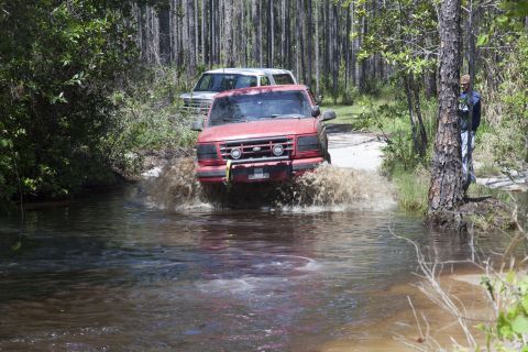 1995 Ford Bronco - Two trucks navigate a water crossing in a wooded area.
