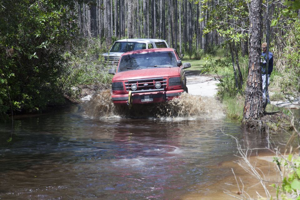 Red pickup truck splashing through a shallow stream with a white SUV behind it