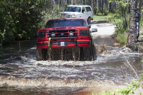 1995 Ford Bronco - Two Ford Broncos navigate a water-filled path in a wooded area.