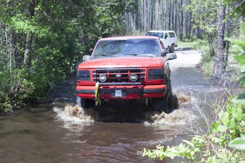 1995 Ford Bronco - A red 1995 Ford Bronco navigates through a water crossing on a dirt road, with another SUV following behind.