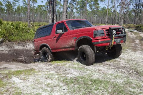 1995 Ford Bronco - This red Ford Bronco is navigating a challenging off-road course with muddy terrain and sandy patches.