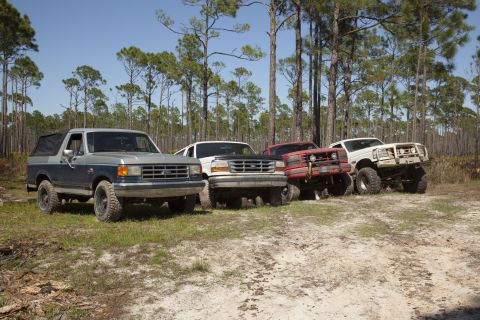 1995 Ford Bronco - A group of modified trucks and SUVs, including a 1987 Ford Bronco, are parked off-road in a forest clearing.