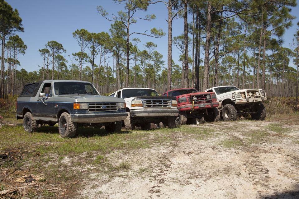Blue and silver 1987 Ford Bronco parked in a wooded area with three other trucks