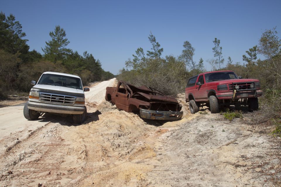 White Ford truck, wrecked rusty truck, and red Ford Bronco on a dirt path