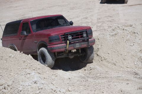 1995 Ford Bronco - A red Ford Bronco navigates a sandy, off-road terrain with another vehicle partially visible in the background.