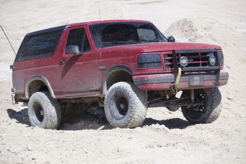 1995 Ford Bronco - This red 1995 Ford Bronco is captured in action, navigating a sandy terrain with its lifted suspension and off-road tires.