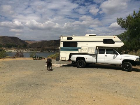 1997 Dodge 3500 - A white truck camper sits parked near a body of water with hills in the background and a dog in the foreground.