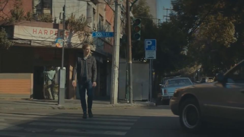Man walks across crosswalk on city street, car in foreground, another car parked on side street