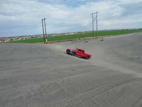 2005 Jeep Wrangler - A red pickup truck navigates a sandy incline, with ATVs visible further up the dune under a cloudy sky.