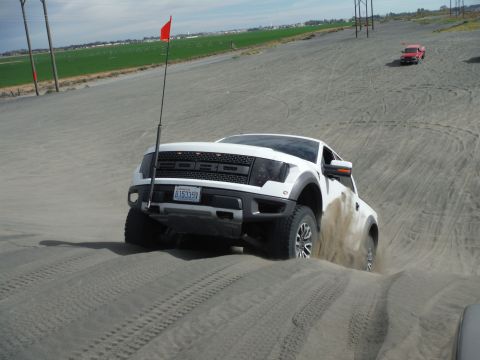 2005 Jeep Wrangler - A white Ford Raptor truck kicks up sand as it navigates a dune, with a smaller red truck visible in the background.