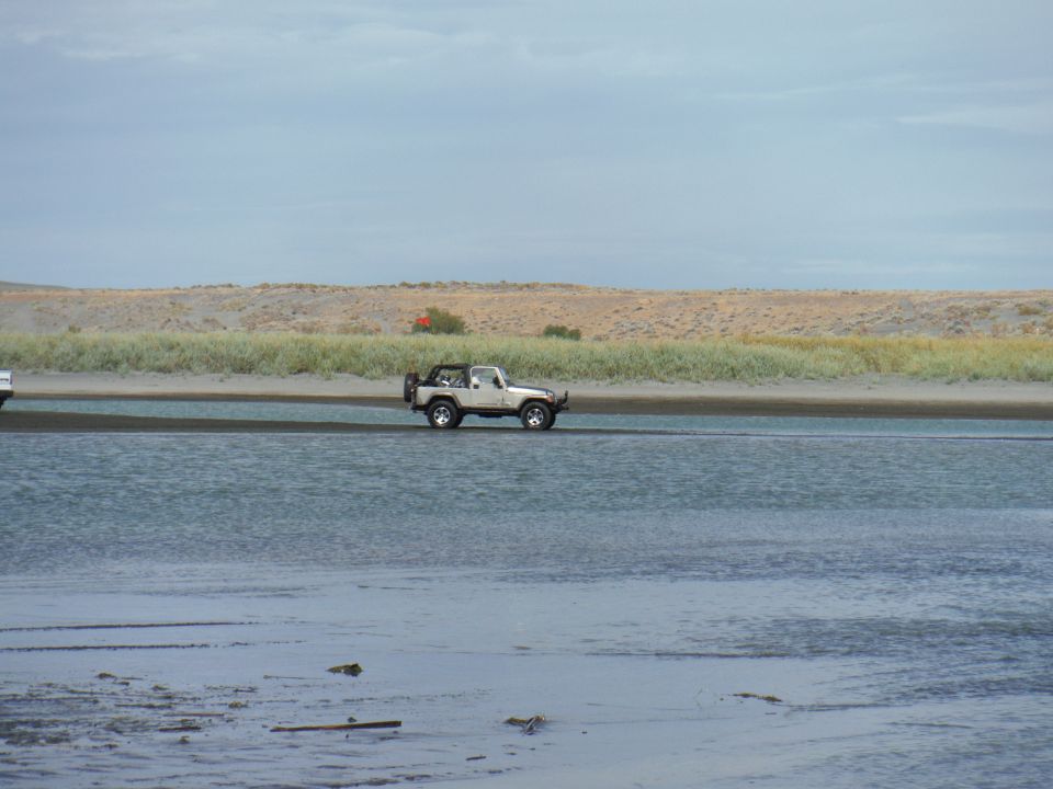 White 2005 Jeep Wrangler driving through shallow water with sandy banks and grass in background