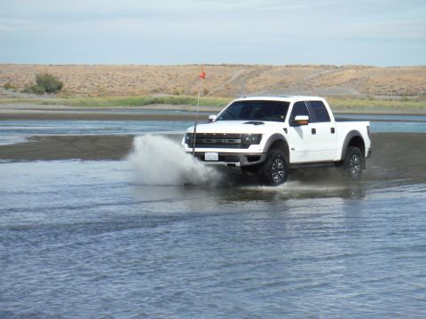 2005 Jeep Wrangler - A white Ford F-150 Raptor truck drives through water, creating a large splash. The truck has a red flag on a pole.