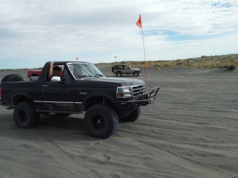2005 Jeep Wrangler - A black 1996 Ford Bronco with a custom bumper and spare tire drives on a sandy terrain with another vehicle in the background.