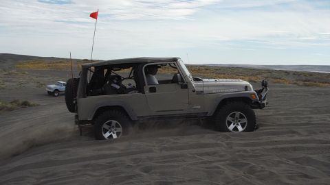 2005 Jeep Wrangler - A Jeep Wrangler navigates sandy terrain, kicking up dust as it goes, with another vehicle in the distance.