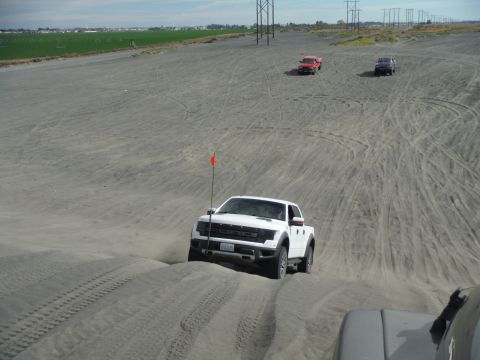 2005 Jeep Wrangler - A white Ford Raptor pickup truck is driving up a sand dune with a red flag attached to a pole on the front fender. Two other pickup trucks…