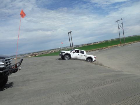 2005 Jeep Wrangler - A white Ford F-150 Raptor pickup truck is driving down a sand dune, kicking up dust. An orange flag is visible on a pole in the foreground.