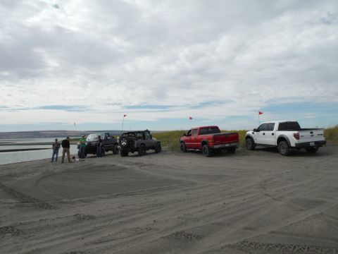 2005 Jeep Wrangler - A group of people stand near several off-road vehicles parked on a sandy shore next to a body of water. The vehicles include a red pickup t…