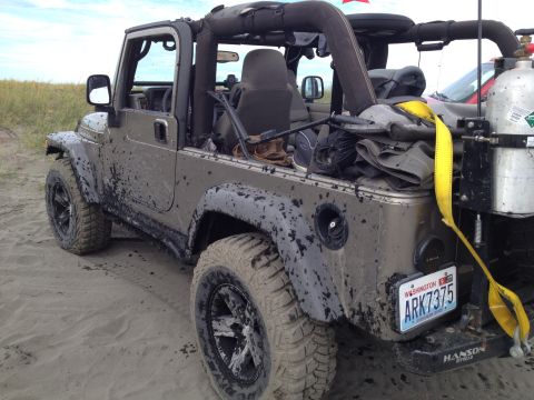 2005 Jeep Wrangler - A mud-splattered, tan Jeep Wrangler is parked on a sandy surface with dry grass in the background. The vehicle has its top off and a yellow…