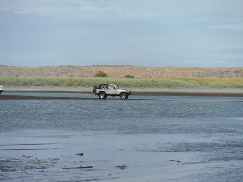 2005 Jeep Wrangler - A white Jeep Wrangler is driving through shallow water on a sandy beach with dunes and vegetation in the background.