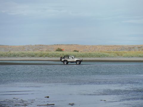 2005 Jeep Wrangler - A white Jeep Wrangler is driving through shallow water with a sandy, grassy embankment in the background under a cloudy sky.