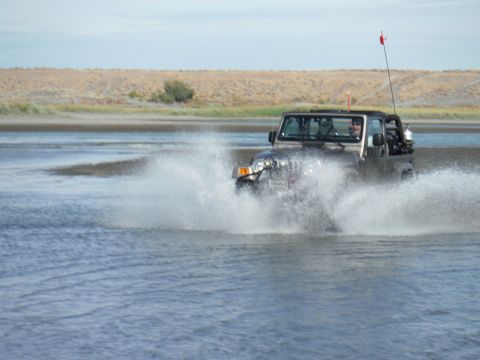 2005 Jeep Wrangler - A silver Jeep Wrangler is driving through shallow water, creating a large splash as it moves forward. The vehicle has a red flag mounted on…