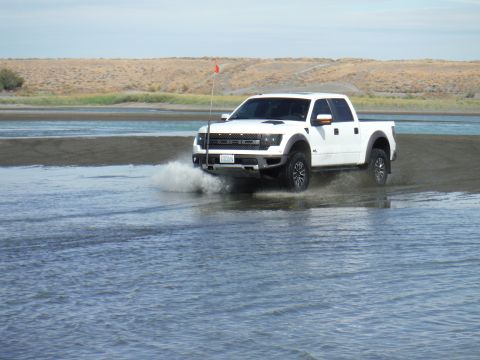 2005 Jeep Wrangler - A white Ford F-150 Raptor pickup truck drives through shallow water, kicking up a spray. The truck has an orange flag mounted on a whip ant…
