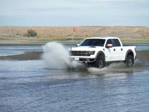 2005 Jeep Wrangler - A white Ford F-150 Raptor pickup truck drives through shallow water, creating a large splash. The truck has an orange flag attached to a wh…