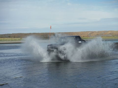 2005 Jeep Wrangler - A dark colored truck drives through shallow water, creating a large splash. A bright orange flag is visible on a pole attached to the truck.