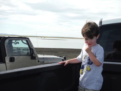 2005 Jeep Wrangler - A young boy stands in the bed of a pickup truck, looking down and touching his finger to his mouth. A tan Jeep Wrangler is visible to the l…