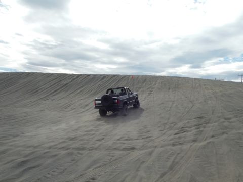2005 Jeep Wrangler - A dark-colored pickup truck is driving up a sandy dune, kicking up dust. The truck has a spare tire mounted on the back.