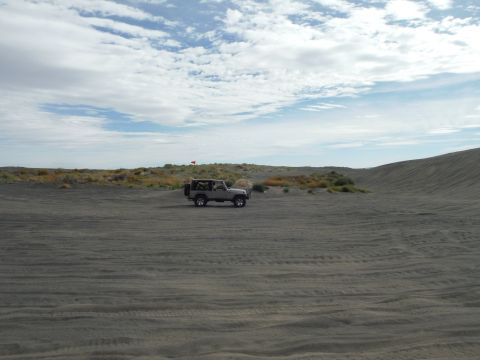 2005 Jeep Wrangler - A gray Jeep Wrangler is parked on a sandy terrain with tire tracks visible in the foreground. A red flag is attached to the spare tire on t…