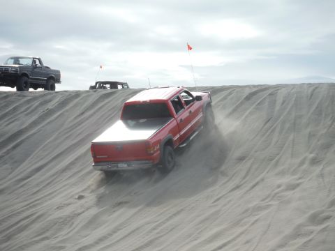 2005 Jeep Wrangler - A red four-door pickup truck with a bed cover drives up a sandy dune, kicking up dust. Another black pickup truck and a dark-colored off-ro…