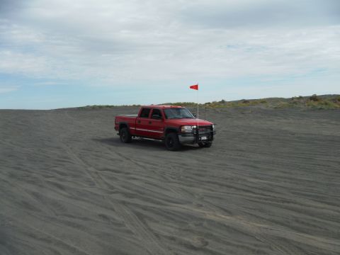 2005 Jeep Wrangler - A red GMC pickup truck with a red flag mounted on the front is parked on a sandy dune under a cloudy sky.