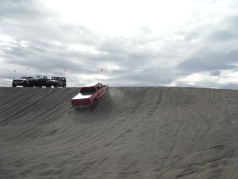 2005 Jeep Wrangler - A red pickup truck is driving down a sand dune, kicking up dust. Several other trucks are parked on the ridge above.