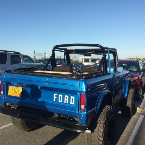 1974 Ford Bronco - The rear driver's side of a bright blue 1974 Ford Bronco is shown parked in a lot under a clear blue sky. The vehicle has a black roll cage…
