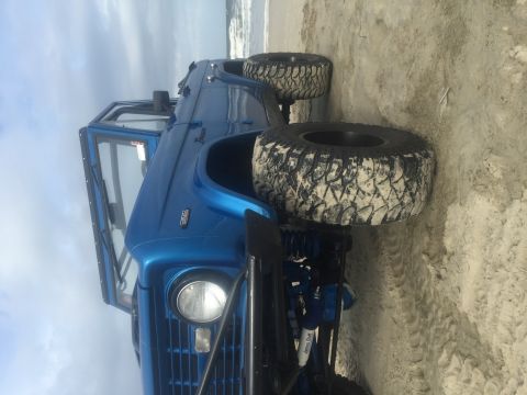 1974 Ford Bronco - A bright blue Ford Bronco is parked on a sandy beach with the ocean in the background. The vehicle has large, aggressive tires covered in s…