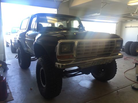 1979 Ford Bronco - A lifted, dark-colored Ford Bronco is parked in a garage with the front end facing the camera. The vehicle has large off-road tires and a c…