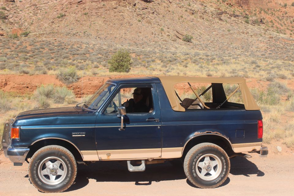 1980s Ford Bronco with tan soft top driving on a dirt road in a desert.