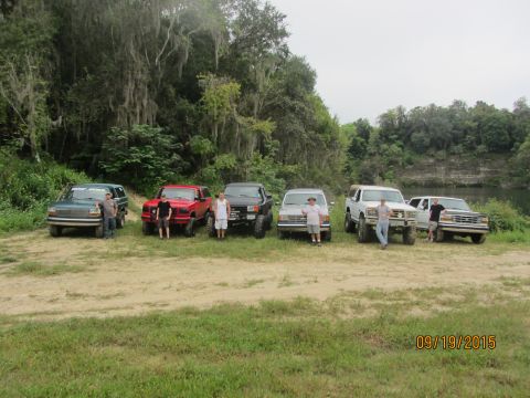 1995 Ford Bronco - Five Ford trucks and SUVs are parked in a line on a dirt path with a wooded area and a body of water in the background. Several men stand n…