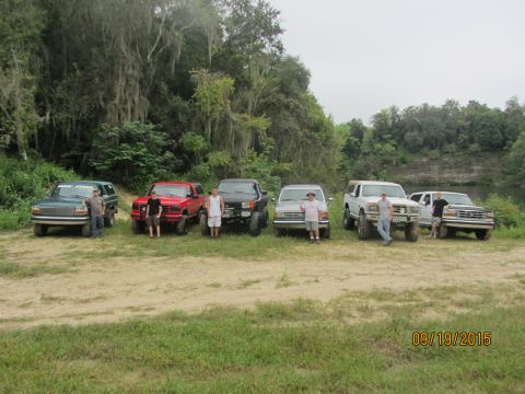 1995 Ford Bronco - Six Ford trucks and SUVs are parked in a line on a dirt path with a wooded area and a river in the background. Five men are standing next t…