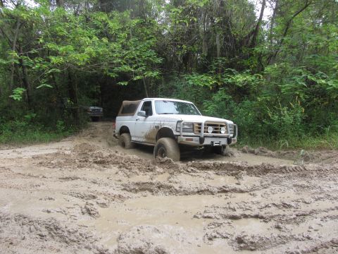 1995 Ford Bronco - A white Ford Bronco is stuck in deep mud on a wooded trail. Another vehicle is visible in the background.