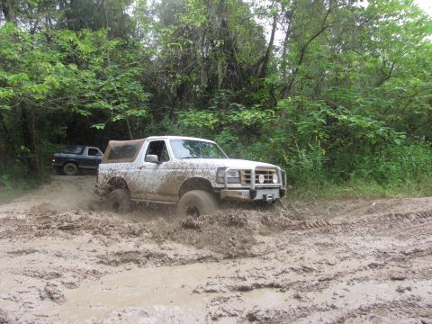 1995 Ford Bronco - A white Ford Bronco is driving through deep mud on a dirt track surrounded by trees. Another dark-colored pickup truck is visible behind it.