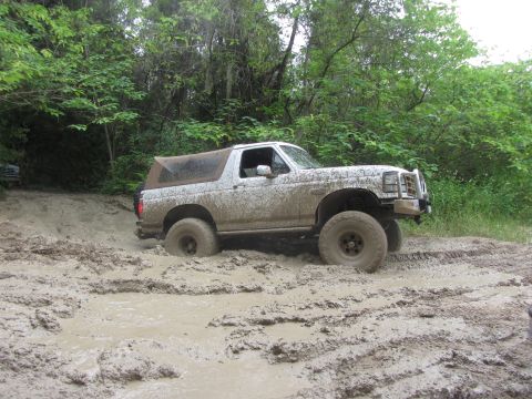 1995 Ford Bronco - A white Ford Bronco is driving through deep mud on a wooded trail. The vehicle is covered in mud and the tires are sunk into the mire.