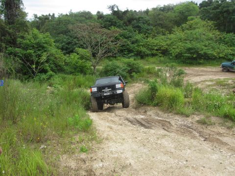 1995 Ford Bronco - A black Ford Bronco is driving on a dirt road surrounded by lush green trees and vegetation. Another green vehicle is visible in the backgr…